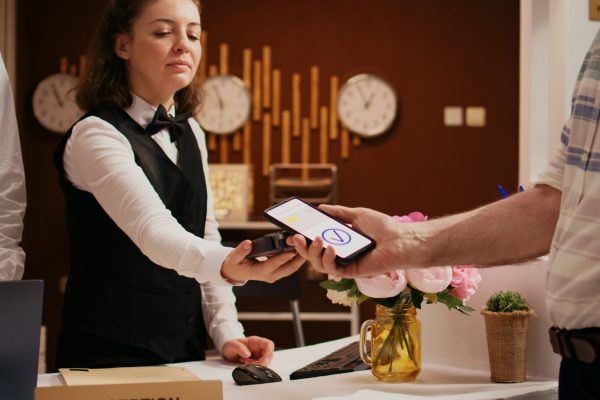 Retired guest using mobile payment with nfc technology at pos terminal, paying for hotel room at reception front desk. Old man registering at holiday retreat, making electronic transaction.
